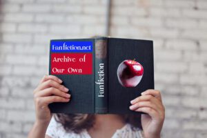 a photo of a female-presenting person holding a black book in front of their face, the spine of the book has been edited to read "Fanfiction", on the back it says "Fanfiction.net" and "Archive of Our Own", on the front a shiny red apple has been edited in.
